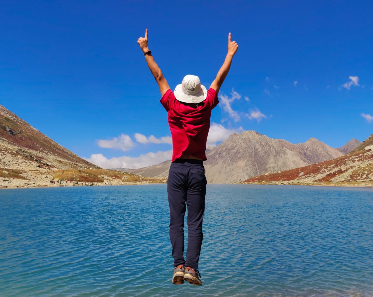 A man in a hat jumps with arms raised near a scenic mountain lake under a clear blue sky.