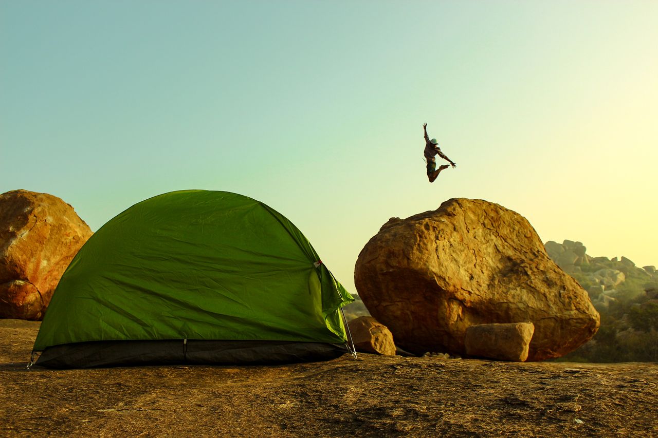Dynamic camping scene with a person jumping near a tent amidst Hampi's rocky landscape.